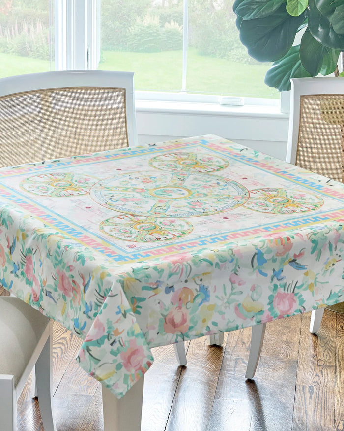 A colorful, floral patterned mahjong tablecloth laid out on a table, with chairs around it and a bright window in the background.