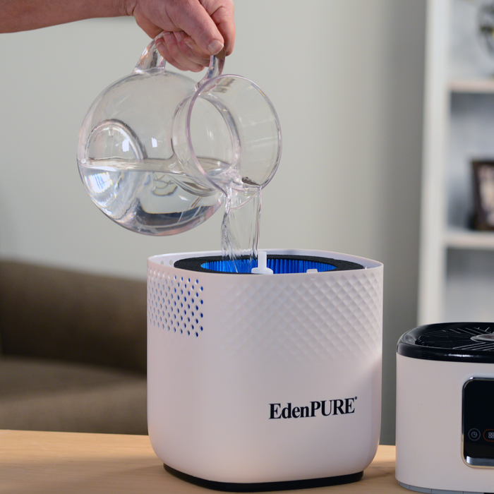 EdenPURE Mist-Free Humidifier with white textured body being filled with water from clear pitcher on wooden table