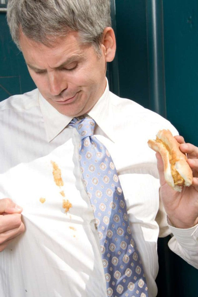 Man in white shirt with blue patterned tie looking at food stain on front of shirt while holding sandwich